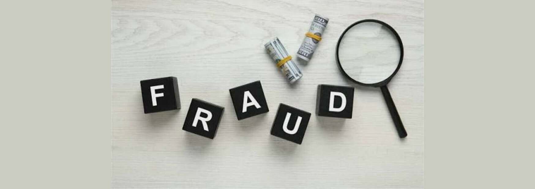 cubi neri che compongono la parola fraud accanto a banconote arrotolate e una lente d'ingrandimento su uno sfondo di legno chiaro.