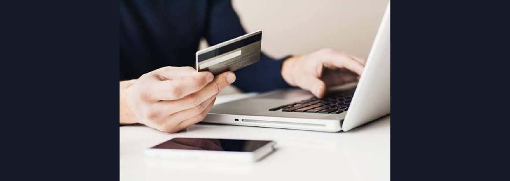 primo piano delle mani di una persona che inserisce i dati di una carta di credito su un computer portatile, con uno smartphone appoggiato sulla scrivania.